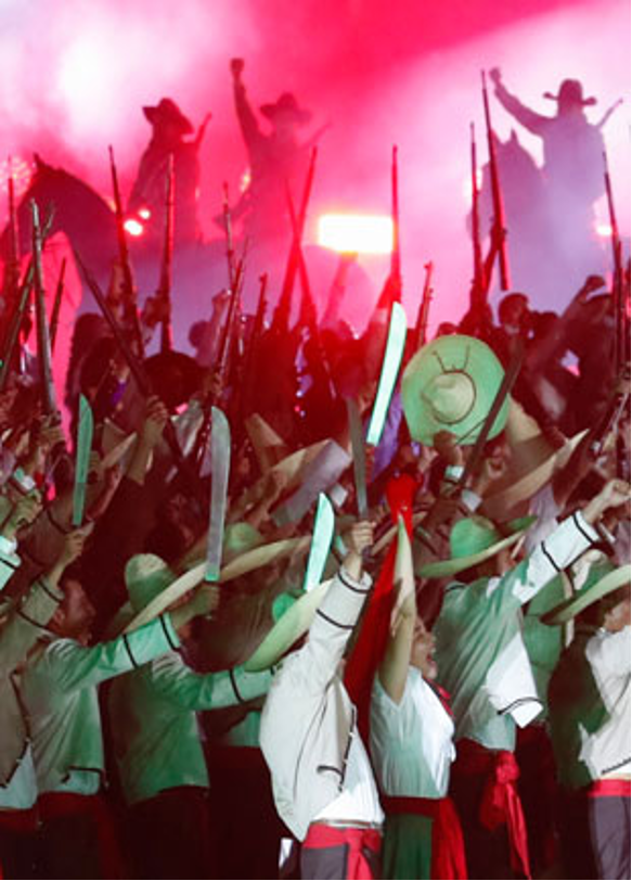 Un grupo de artistas vestidos de rojo y verde levantan machetes y saludan bajo un escenario iluminado en rojo, con siluetas en el fondo.