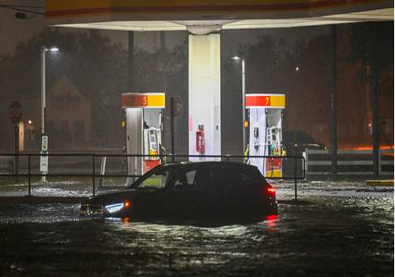 Un automóvil está parcialmente sumergido bajo el agua de la inundación en una gasolinera durante la noche, con dos bombas de combustible en el fondo.