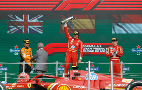 Tres pilotos de carreras se encuentran de pie en un podio, con la figura central sosteniendo un trofeo en alto. Detrás de ellos se exhiben las banderas del Reino Unido, España y México.