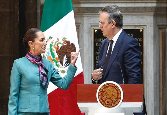 Dos personas vestidas de gala conversan cerca de un podio y una bandera mexicana. Una de ellas hace un gesto con el dedo en alto.