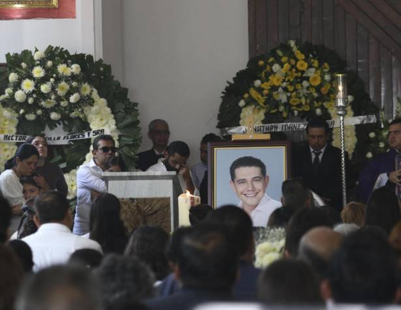 Un servicio conmemorativo con los asistentes reunidos alrededor de un retrato de un hombre sonriente, rodeado de arreglos florales y velas encendidas.