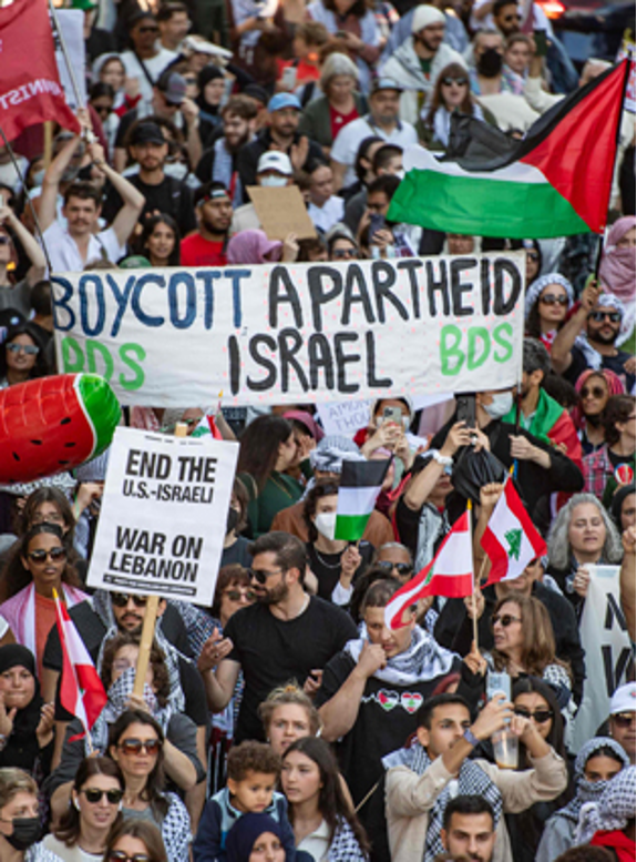 Una gran multitud se reúne en una protesta, sosteniendo carteles que dicen "Boicot al Apartheid de Israel", "Poner fin a la guerra entre Estados Unidos e Israel en el Líbano" y se ven varias banderas palestinas.