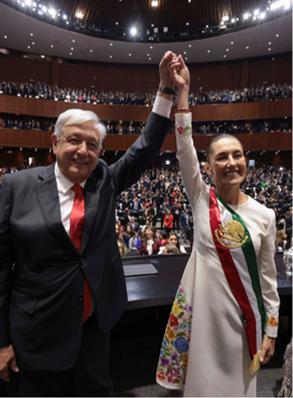 Dos personas, una con traje y corbata roja y la otra con un vestido blanco con faja tricolor, se toman de la mano en alto en un gran auditorio lleno de personas sentadas.