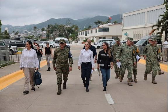 Un grupo de personas, entre militares y civiles, caminan juntos al aire libre cerca de un edificio con montañas al fondo.