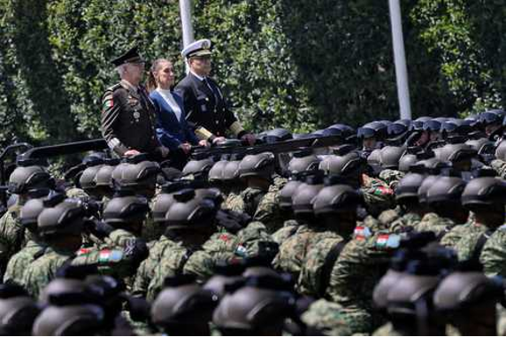 Oficiales militares uniformados viajan en un vehículo abierto junto a una fila de soldados que visten cascos y uniformes de camuflaje.