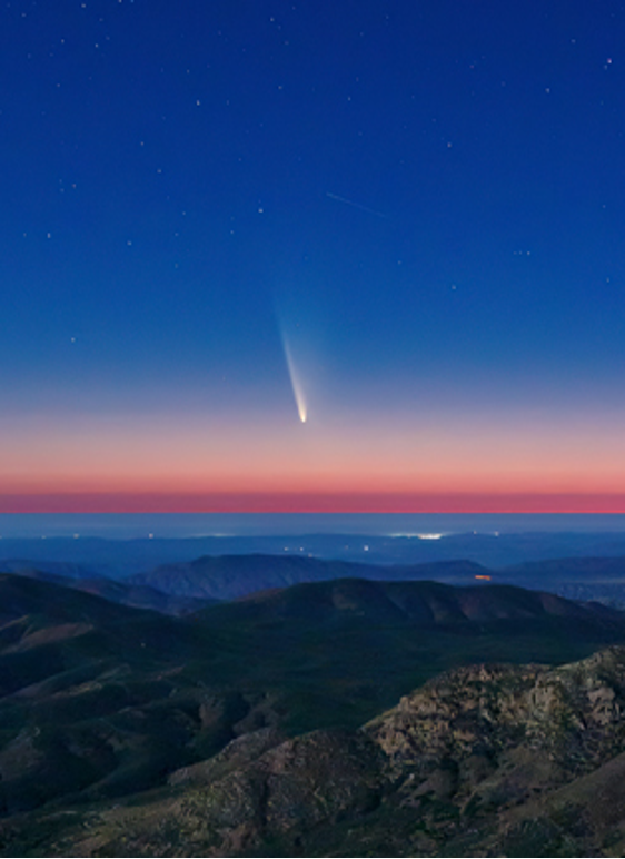 Un cometa con una cola brillante brilla en el cielo crepuscular sobre una cadena montañosa.