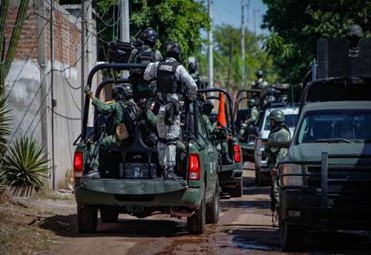 Personal militar con equipo táctico viajando dentro de camiones que recorren una calle estrecha y fangosa bordeada de árboles y edificios.