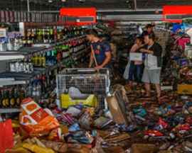 Las personas recorren un pasillo desordenado en una tienda con artículos dispersos y escombros en el piso. Los estantes están desorganizados y una persona empuja un carrito mientras otras llevan artículos.