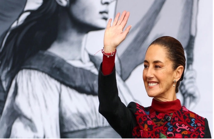 Una persona saludando, sonriendo, frente a un gran mural monocromático que representa figuras.