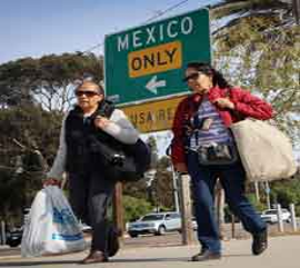 Dos personas pasan junto a un cartel que indica la dirección a México, cargando bolsas. Una de ellas lleva gafas de sol y un chaleco negro, mientras que la otra lleva una chaqueta roja. Se ven árboles al fondo.