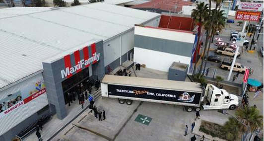 Un camión con el mensaje "Tastes Like Miller Time, California" está estacionado cerca de la entrada de una tienda MaxFamily. Hay gente reunida afuera.