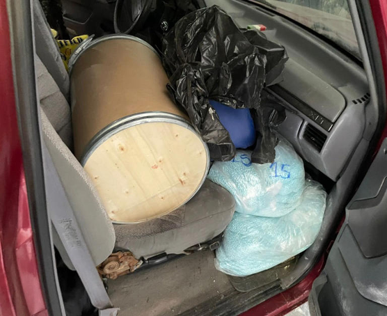 Interior del automóvil con barril de madera en el asiento trasero, bolsas de plástico negras y bolsas azules grandes etiquetadas con texto blanco.