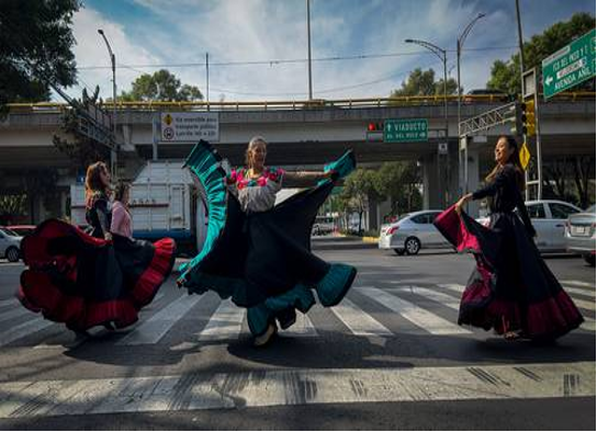 Tres personas con trajes tradicionales bailan en un cruce de peatones de la ciudad con coches y un paso elevado al fondo.