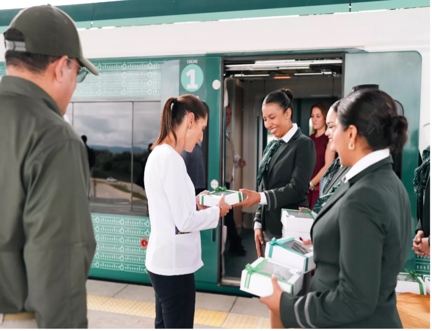 Varias personas se encuentran de pie junto a un tren intercambiando cajas envueltas para regalo. Una persona con camisa blanca acepta una de un hombre uniformado. Otras dos personas observan, vestidas con uniformes similares.