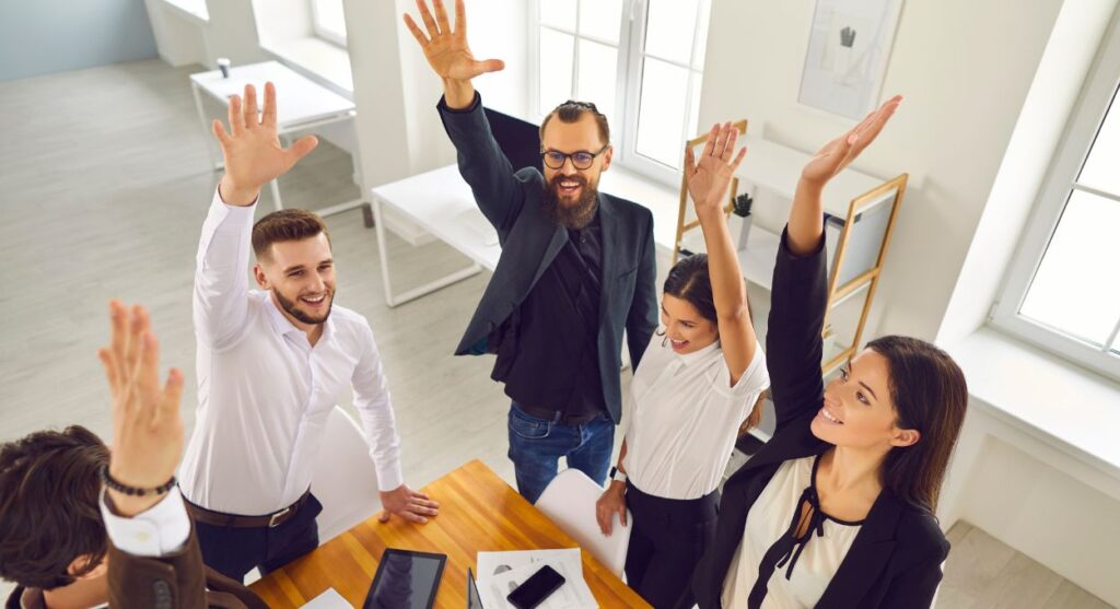 Cinco personas vestidas de negocios están de pie alrededor de una mesa con las manos levantadas, pareciendo entusiastas en un entorno de oficina luminoso, ejemplificando el trabajo en equipo.