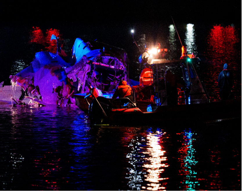 Los equipos de rescate inspeccionan un helicóptero dañado en el agua por la noche, iluminado por luces de emergencia que reflejan colores vibrantes en la superficie.