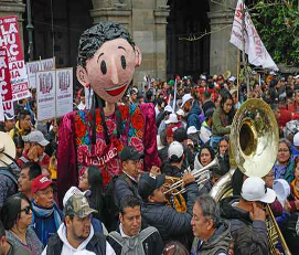 Multitud en un animado evento al aire libre con una gran marioneta vestida de colores y una banda de música.