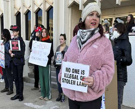 Un grupo de personas se encuentra de pie en unas escaleras con carteles que incluyen mensajes sobre inmigración y apoyo a los niños. Uno de los carteles dice: "Nadie es ilegal en tierras robadas".