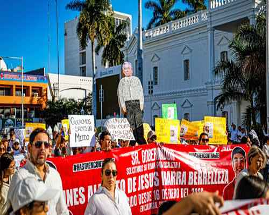 Una multitud de personas con carteles y pancartas marcha en una protesta. Se ve una silueta de una persona sobre la multitud. El fondo muestra un edificio blanco y palmeras.