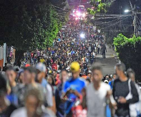 Una gran multitud de personas caminando por una calle poco iluminada y bordeada de árboles, con luces rojas de vehículos visibles en el fondo.