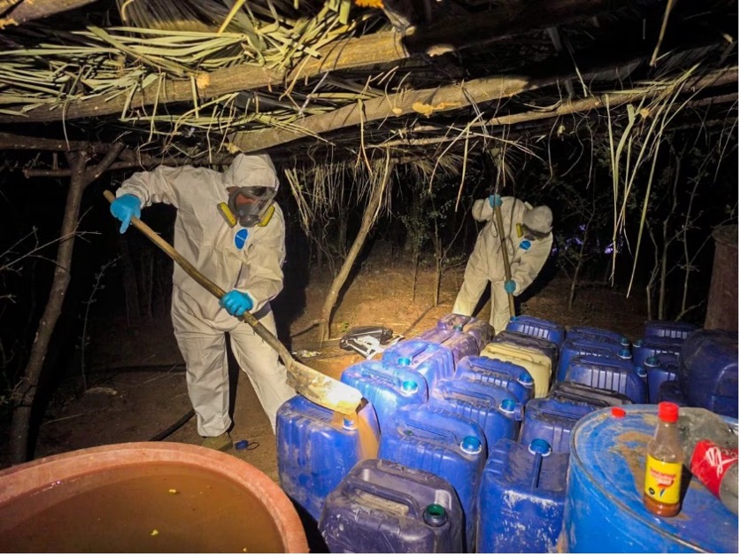 Dos personas con trajes protectores y máscaras trabajan con contenedores azules bajo una estructura con techo de paja durante la noche.