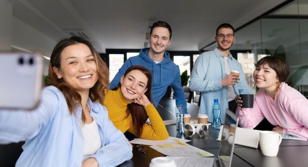 Cinco personas sonriendo y tomándose un selfie en una sala de conferencias, rodeadas de papeles y bebidas en la mesa, celebrando el empoderamiento de las nuevas generaciones.