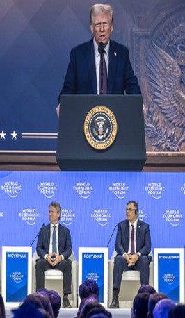 Un orador se encuentra en un podio con el sello presidencial, mientras tres personas se sientan en un escenario con carteles del Foro Económico Mundial detrás de ellas.