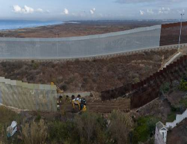 Vista aérea de maquinaria de construcción trabajando en una valla fronteriza en un paisaje desértico, con la costa visible al fondo.