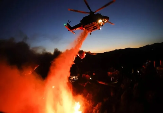 Un helicóptero arroja agua sobre un gran incendio en el suelo contra un cielo crepuscular, con el paisaje circundante parcialmente visible.