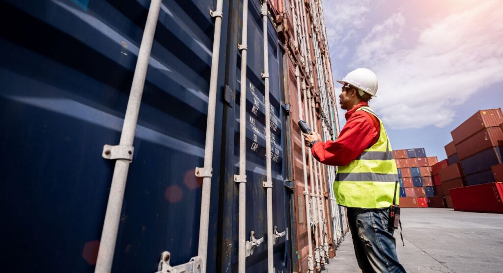 Un trabajador con equipo de seguridad inspecciona meticulosamente los contenedores de envío apilados al aire libre, asegurándose de que cada detalle se alinee con los estándares de precisión de La Exportación.