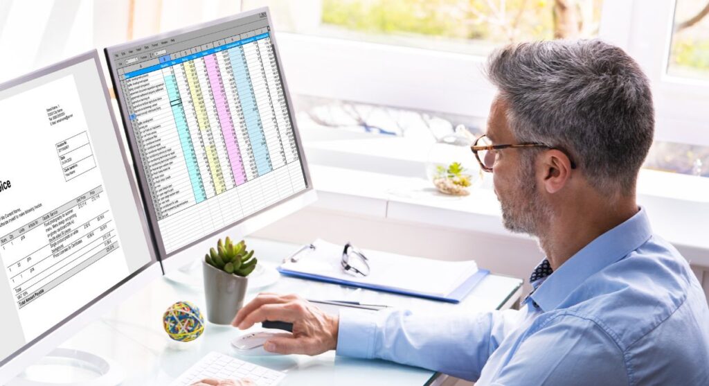 Un hombre con camisa azul trabaja en un escritorio con dos monitores que muestran hojas de cálculo y analiza las tendencias en la fiscalización. Una planta y una bola decorativa añaden un toque de naturaleza y creatividad a su espacio de trabajo.