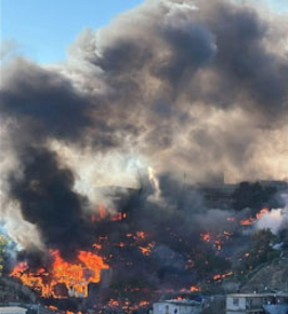 Un humo negro espeso y grandes llamas se elevan desde un incendio en la ladera de una colina, con algunas estructuras visibles en la parte inferior.