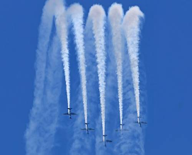 Seis aviones volando en formación emiten estelas de humo blanco contra un cielo azul claro.