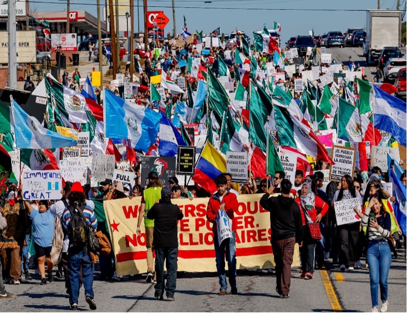 Una gran multitud sostiene varias banderas y carteles de protesta mientras marcha por una calle.