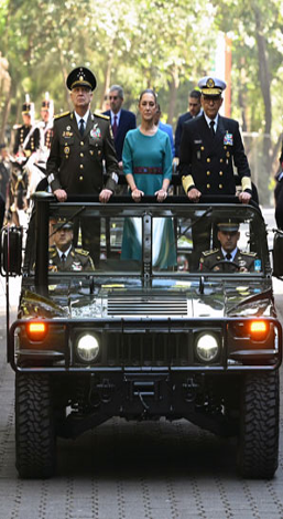 Tres personas vestidas con uniforme militar formal se encuentran en un vehículo de techo abierto en movimiento, con soldados en el fondo y árboles que bordean un camino iluminado por el sol.