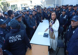 Una mujer habla a través de un micrófono mientras está de pie en una mesa rodeada de oficiales de policía uniformados en una reunión al aire libre.