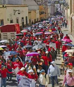 Un gran grupo de personas marcha por la calle, algunas con paraguas y carteles. Muchas llevan camisetas rojas. Los edificios bordean la calle a ambos lados.