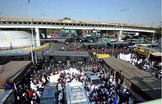 Una gran multitud se reúne cerca de un mercado callejero, con vehículos policiales y oficiales formando una barricada bajo un paso elevado.