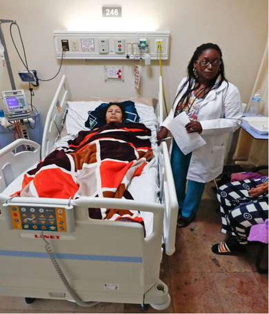 Un médico se encuentra junto a una mujer acostada en una cama de hospital, cubierta con una manta estampada. Alrededor de la cama hay equipo médico.