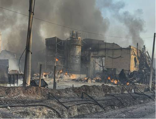 Sitio industrial con graves daños por incendio, humo saliendo de los edificios, escombros esparcidos y pequeños incendios aún activos.