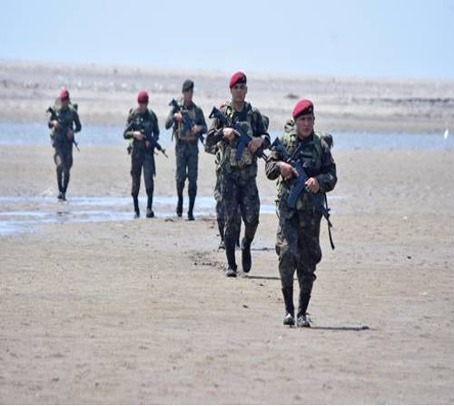 Cinco soldados con uniformes de camuflaje y boinas rojas caminan en formación sobre un terreno arenoso, portando rifles.