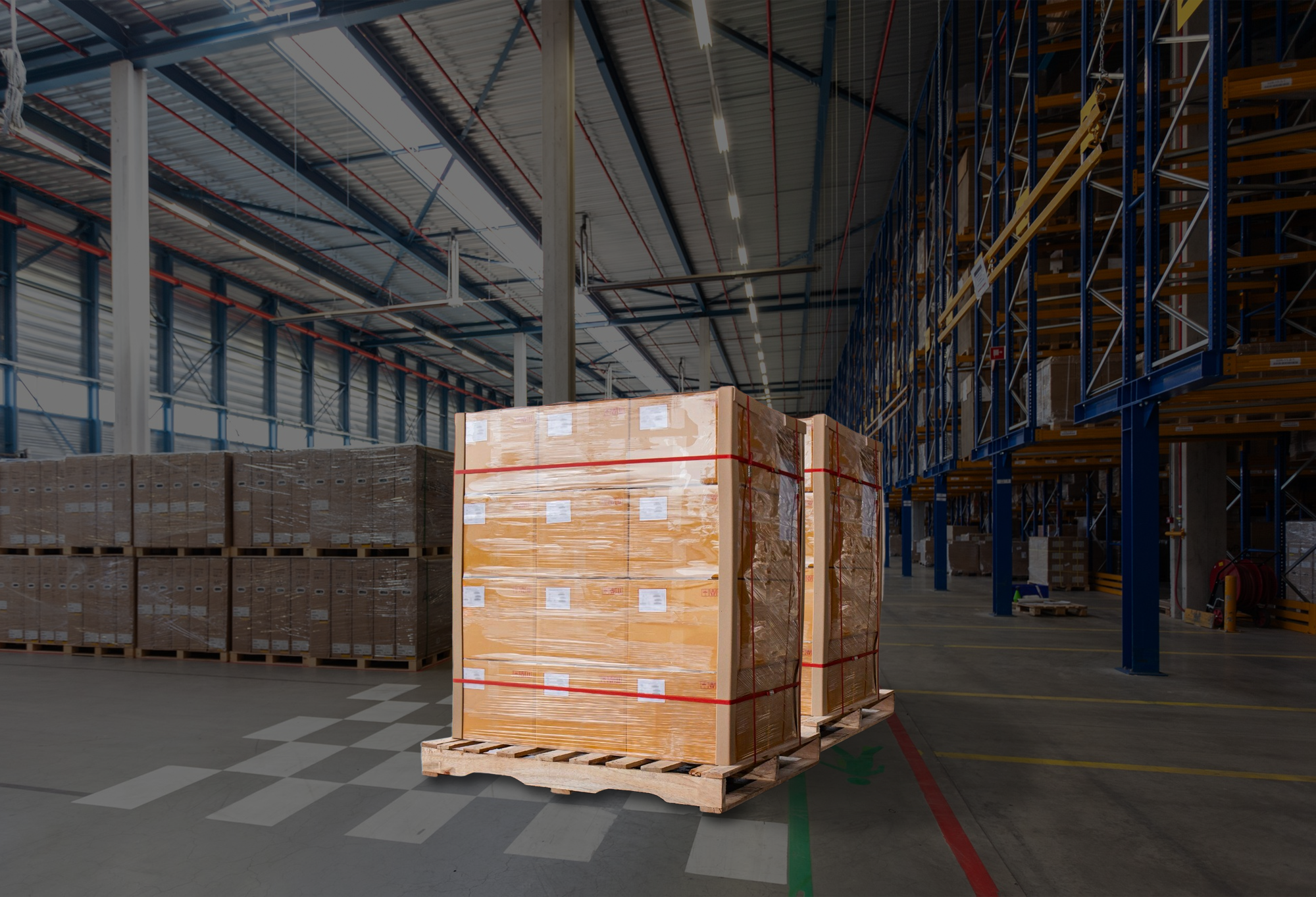En el suelo de un almacén se coloca un palé de madera cargado con cajas bien envueltas. Al fondo se ven varios estantes y pilas de palets similares.