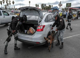 Agentes de policía y un perro inspeccionan el maletero abierto de un coche blanco en una calle muy transitada con varios vehículos y personas al fondo.