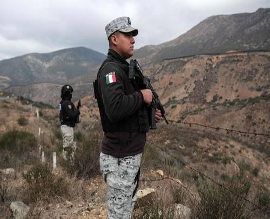 Dos agentes uniformados se encuentran junto a una valla en una zona montañosa, uno de ellos con un fusil en la mano. Parecen estar vigilando la región.