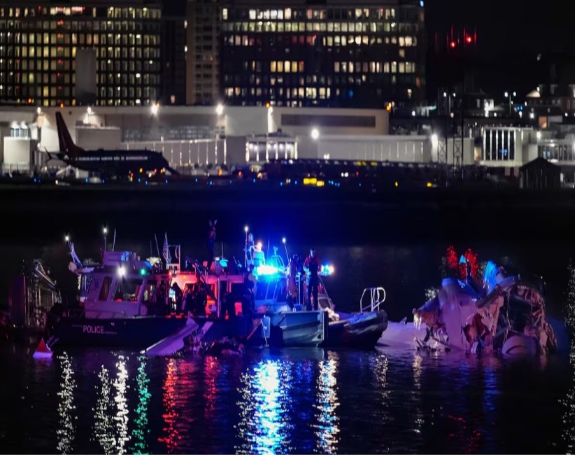 Policía y barcos de rescate por la noche en una vía fluvial cerca de un avión con daños visibles, luces de la ciudad y el aeropuerto al fondo.