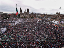 Una gran multitud se reúne en una plaza pública con una bandera mexicana, rodeada de edificios históricos bajo un cielo nublado.