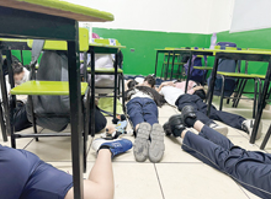 Estudiantes acostados en el suelo debajo de los escritorios en un aula con paredes verdes y azulejos beige, posiblemente durante un simulacro o una situación de emergencia.