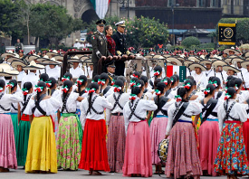Un desfile militar con soldados vestidos con trajes tradicionales y coloridos marchando, liderados por oficiales uniformados en una plaza de la ciudad.