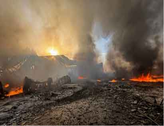 Un gran incendio arde y genera una densa humareda y escombros. Las llamas y el humo envuelven las estructuras bajo un cielo anaranjado.