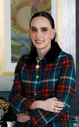 Una persona con cabello largo y oscuro, que viste una chaqueta a cuadros y joyas, estÃ¡ de pie con los brazos cruzados frente a una obra de arte enmarcada.